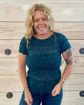 Woman wearing a teal patterned shirt and blue jeans standing against a wooden background