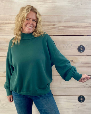 Woman wearing a green sweater and blue jeans standing against a wooden wall.