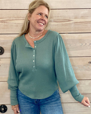 Woman wearing a teal long-sleeve shirt and blue jeans standing against a wooden wall.