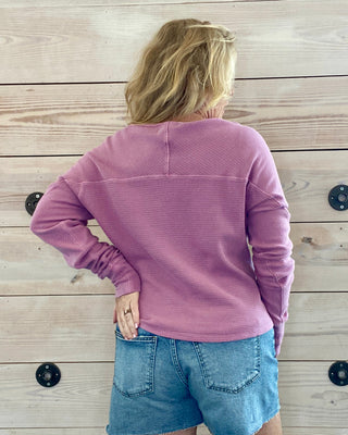 Person wearing a pink long-sleeve shirt and denim shorts standing against a wooden wall.