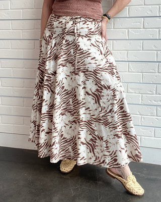 Woman wearing a patterned top and long skirt against a white brick wall.