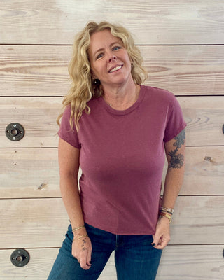 Woman wearing a maroon t-shirt and blue jeans standing against a wooden wall.