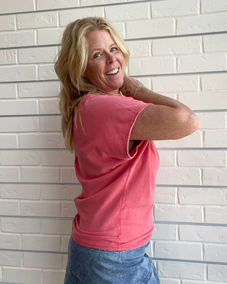 Woman in a pink shirt and blue skirt standing against a light brick wall.