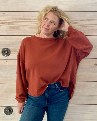 Woman wearing a rust-colored sweater and blue jeans against a wooden wall.