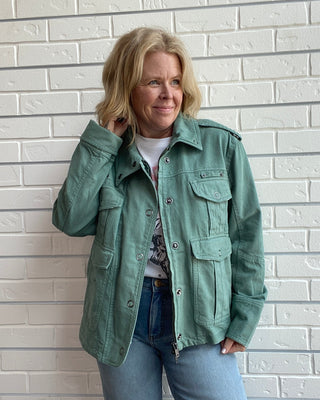 Person wearing a green jacket and blue jeans standing against a light brick wall.