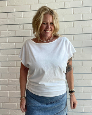 Woman wearing a white t-shirt and blue skirt standing against a light brick wall.