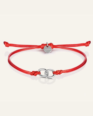 A red adjustable cord bracelet with a polished silver-tone double-ring centerpiece and a small round silver charm, displayed on a light cream background.