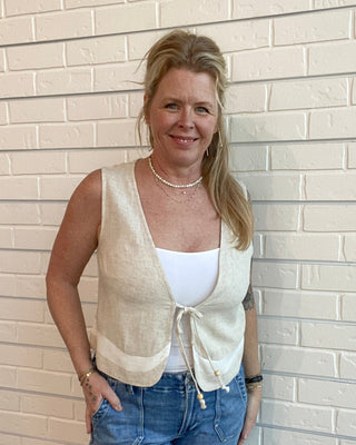 Woman wearing a beige vest over a white top and blue jeans against a light brick wall.