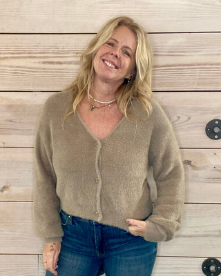 Woman wearing a beige cardigan and blue jeans standing against a wooden wall.