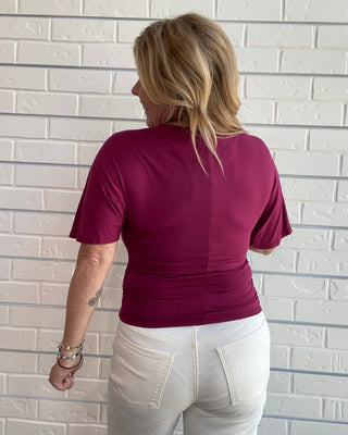 A woman viewed from behind wearing a short-sleeve crewneck t-shirt in deep burgundy and white pants, standing against a white brick wall.