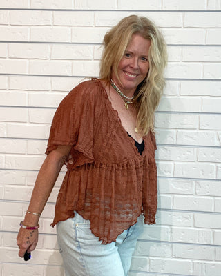 A woman with blonde hair wearing a rust-brown ruffled short-sleeve top and light-wash jeans, standing against a white brick wall.