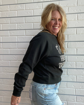 A smiling blonde woman wearing a black crewneck sweatshirt with white graphic text and light blue denim shorts, posing against a white brick wall.