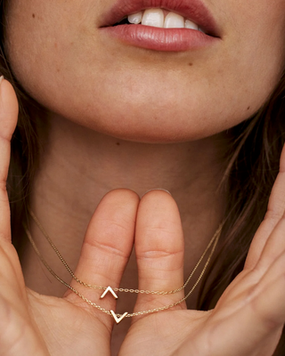 A close-up lifestyle shot of a model wearing delicate gold necklaces with small V-shaped initial pendants, held gently in her hands.