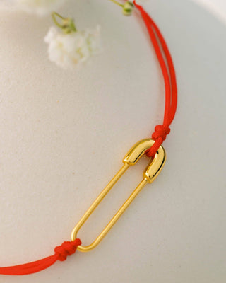 A close-up of a red cord bracelet featuring a polished gold safety-pin style charm, resting on a light beige textured surface with soft white flowers in the background.