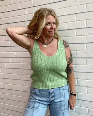 Woman wearing a green sleeveless top and blue jeans standing against a light brick wall.