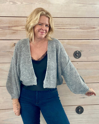 Woman wearing a gray cardigan over a black top and blue jeans against a wooden background
