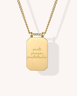 A gold rectangular pendant necklace with a chain, featuring the engraved script text 'grit grace gratitude' on a light cream background.