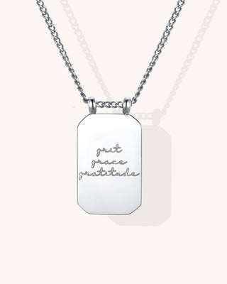 A silver-toned dog tag necklace with a rectangular pendant engraved with the words 'grit grace gratitude' in cursive, shown on a light cream background.