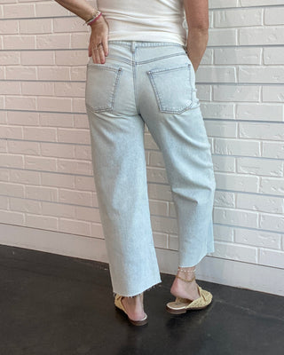 Person wearing a white sleeveless top and light blue jeans against a white brick wall.