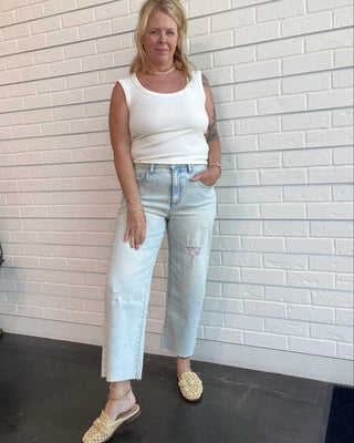 Woman wearing a white sleeveless top and light blue jeans standing against a white brick wall.