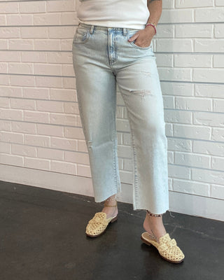 Woman wearing a white tank top and light blue jeans standing against a white brick wall.