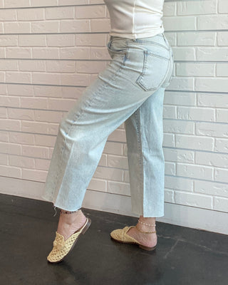 Woman wearing a white tank top and light blue jeans against a white brick wall.