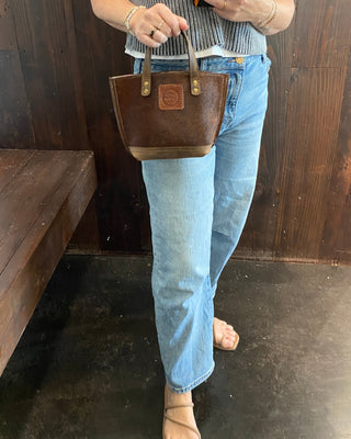 A woman wearing a white sleeveless top and light blue jeans holds a brown leather handbag with gold-tone hardware, standing in front of a dark wood-paneled wall.