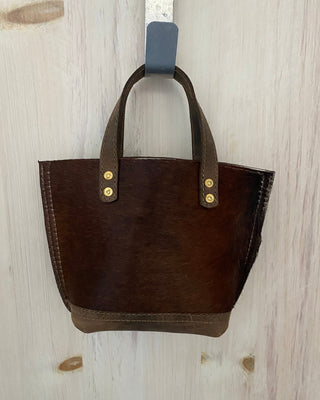 A small dark brown leather tote bag with double top handles and gold-tone rivets, hanging from a gray hook on a light-colored wooden door.