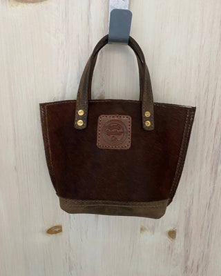 A small dark brown leather tote bag with double top handles and brass-tone rivets, hanging from a gray hook against a light wood-paneled wall.