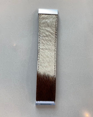 A long rectangular leather strap with a natural cowhide pattern, featuring a white top section and a dark brown bottom section, finished with silver-toned metal hardware on a light gray textured background.