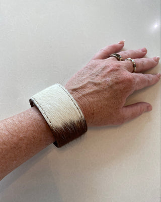 A close-up of a person's forearm and hand wearing a wide cuff bracelet made from cowhide, with a natural white and dark brown gradient and a fringed edge, set against a light beige background.