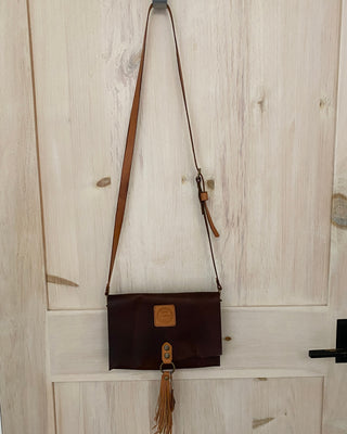 Brown leather crossbody bag with a tassel hanging on a wooden door.