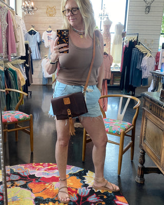 Woman taking a mirror selfie in a clothing store wearing a brown tank top and light blue shorts.
