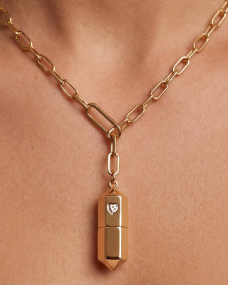 A close-up of a gold chain necklace worn on a person's neck, featuring a bullet-shaped pendant with a small heart-shaped gemstone on the front.