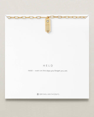 A gold chain necklace with a small rectangular pendant and a tiny clear stone, displayed on a white card that reads 'HELD' with the brand name Bryan Anthony's at the bottom.