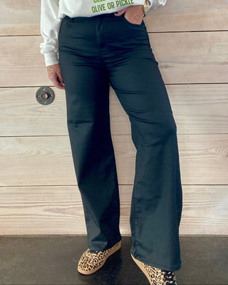 Woman wearing a white sweatshirt with colorful text and dark pants standing against a wooden wall.