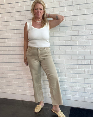Woman wearing a white tank top and beige pants against a white brick wall.