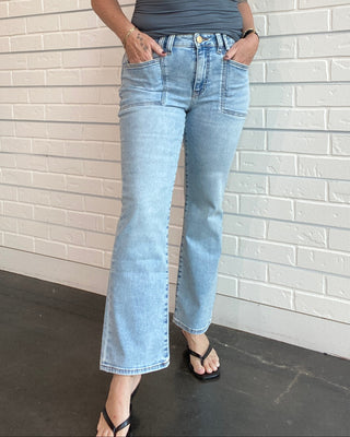 Woman wearing a gray tank top and light blue jeans standing against a white brick wall.