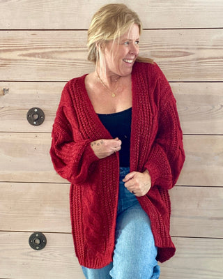 Woman wearing a red cardigan and blue jeans standing against a wooden wall.