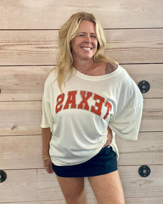 Woman wearing a white 'TEXAS' shirt and dark shorts standing against a wooden wall.