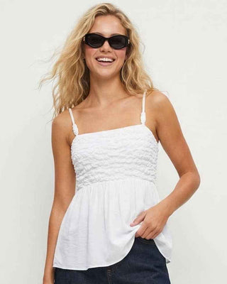 Woman wearing a white top and dark shorts with sunglasses on a plain background