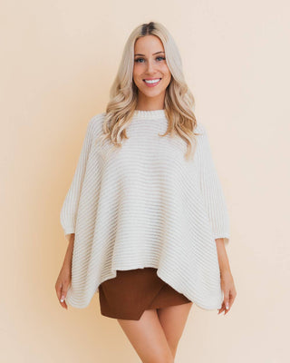 Woman wearing a white poncho over a brown dress with knee-high boots on a beige background