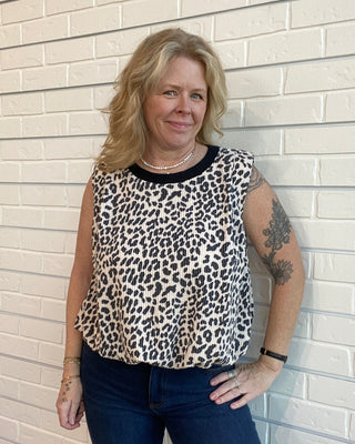 Woman wearing a leopard print top and blue jeans standing against a light brick wall.