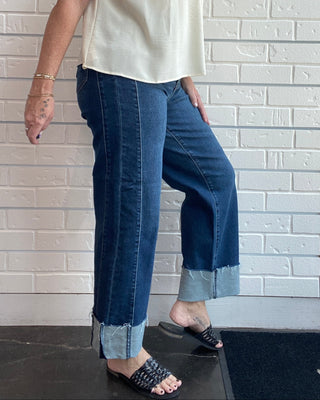 A woman modeling a sleeveless cream top and dark blue cropped jeans with rolled cuffs, standing against a white brick wall.