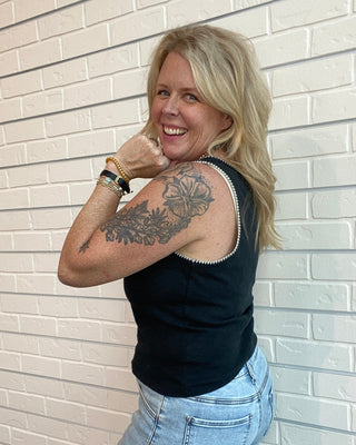 Woman with tattoos on arm and back wearing a black sleeveless top and blue jeans against a light brick wall.