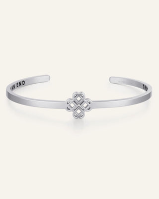 A silver-toned metal cuff bracelet with a central knotted geometric charm and engraved text inside the arms, shown on a plain light beige background.
