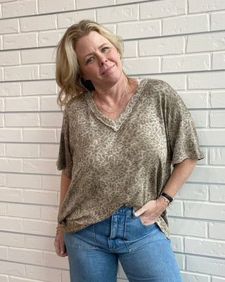 Woman wearing a patterned top and blue jeans standing against a light brick wall.