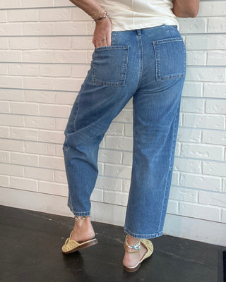 A person shown from the back wearing blue wide-leg cropped jeans, a cream sleeveless top, and beige flat sandals, standing against a white brick wall.
