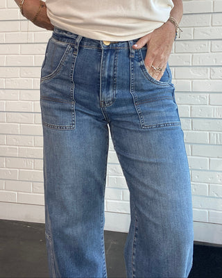 A person wearing blue wide-leg jeans and a cream sleeveless top, standing against a white brick wall.