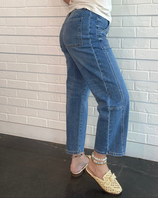 A woman wearing blue cropped denim jeans, a white sleeveless top, and beige woven mule sandals, standing against a white brick wall.